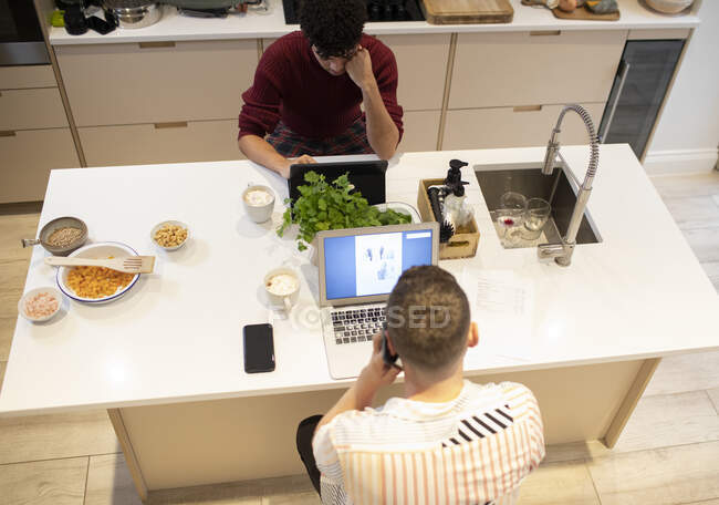 Young gay male couple working from home at laptops in kitchen — Stock Photo