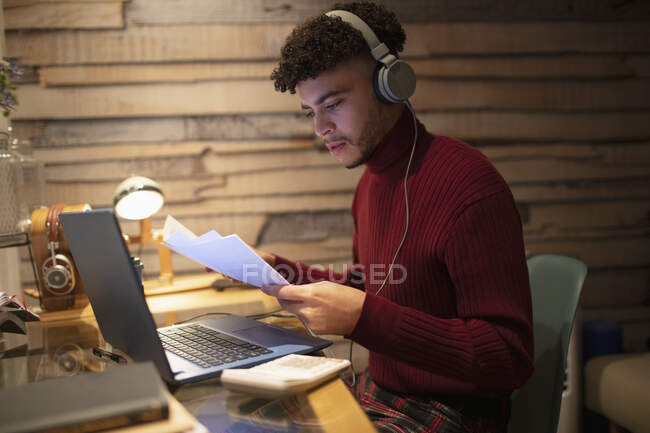 Молодий чоловік з навушниками та паперовими роботами, що працюють пізно вдома — стокове фото
