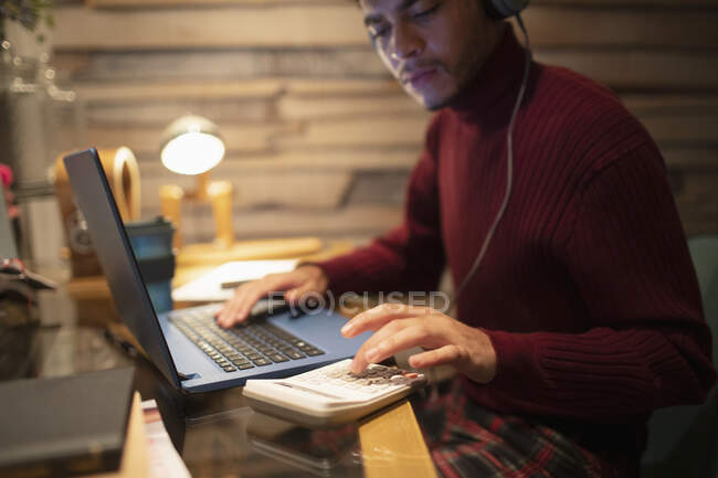 Young man with headphones using calculator at laptop in home office — Stock Photo