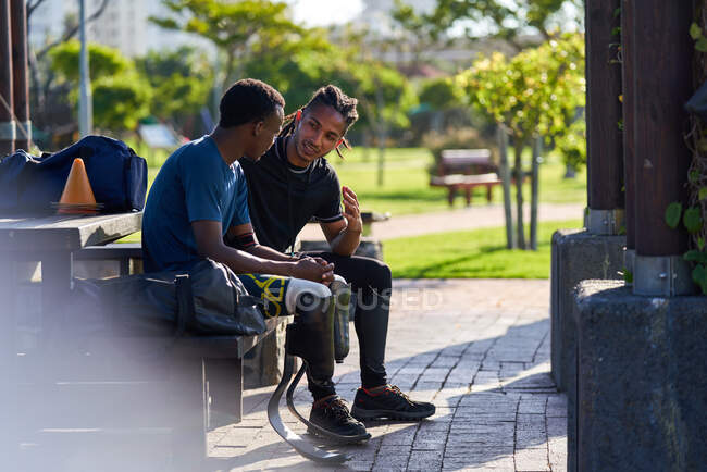 Coach and young male amputee athlete talking on park bench — Stock Photo