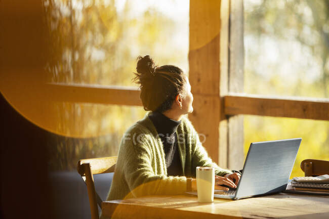 Жінка працює на ноутбуці, дивлячись у вікно в сонячному кафе — стокове фото