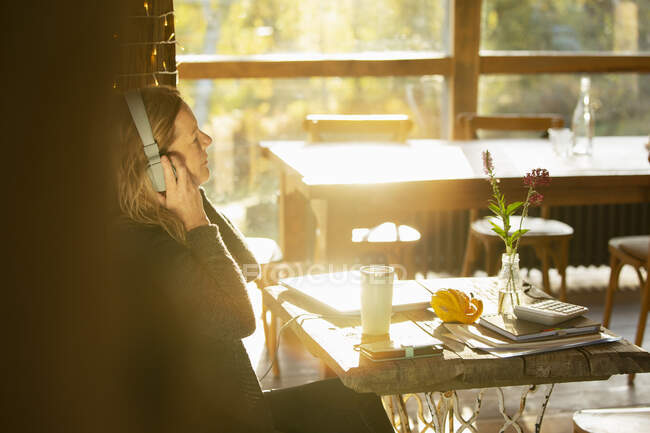 Empresaria con auriculares trabajando en cafetería soleada - foto de stock