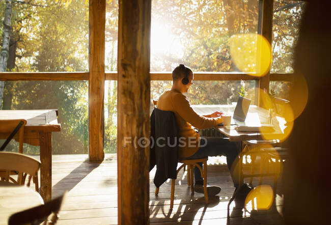 Uomo d'affari con cuffie che lavorano al laptop in un bar soleggiato — Foto stock