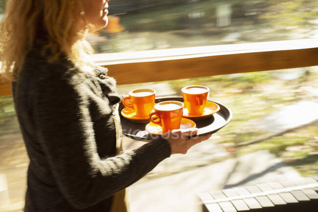 Женщина-бариста несет поднос с кофейными чашками вдоль окна солнечного кафе — стоковое фото