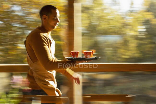 Мужчина-бариста несет поднос с кофейными чашками вдоль осеннего окна кафе — стоковое фото