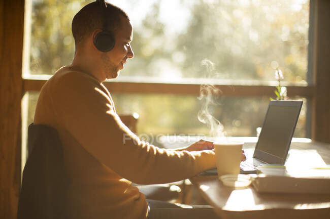 Empresário com fones de ouvido e café quente trabalhando no laptop no café — Fotografia de Stock