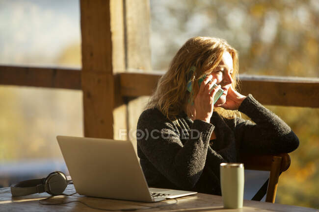 Businesswoman talking on smart phone at laptop in sunny cafe — Stock Photo