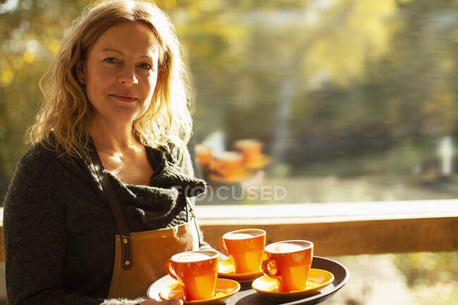 Retrato confiado barista femenina con bandeja de café en ventana soleada - foto de stock
