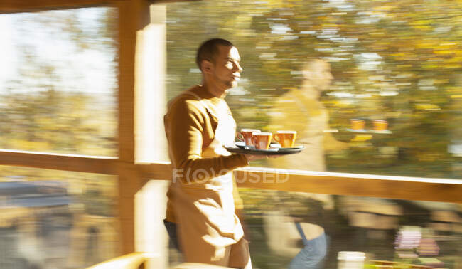 Barista maschio che trasporta vassoio di caffè lungo soleggiata finestra autunnale caffè — Foto stock