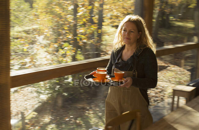 Female barista carrying coffee on tray along sunny autumn cafe window — Stock Photo