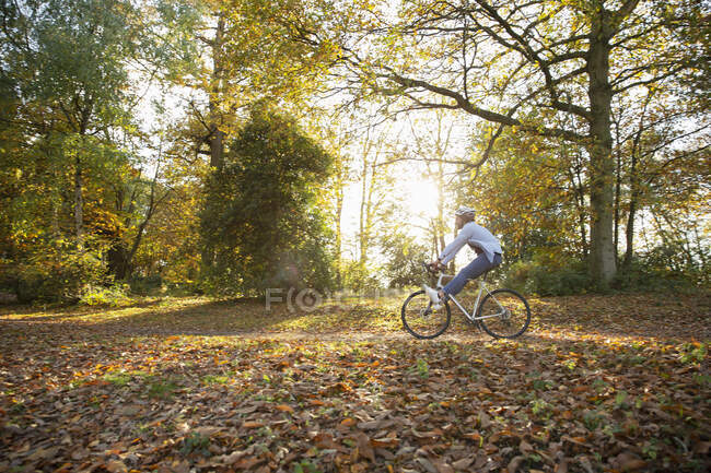 Mujer joven despreocupada montar en bicicleta a través de hojas de otoño en el parque - foto de stock