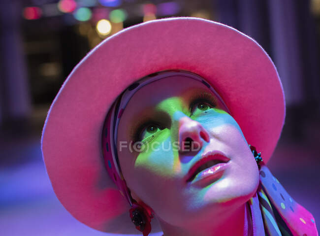 Curiosa mujer con estilo en sombrero rosa mirando hacia arriba en luz de neón - foto de stock