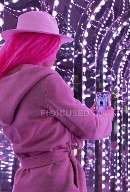 Mulher na moda em casaco rosa usando telefone câmera sob luzes — Fotografia de Stock
