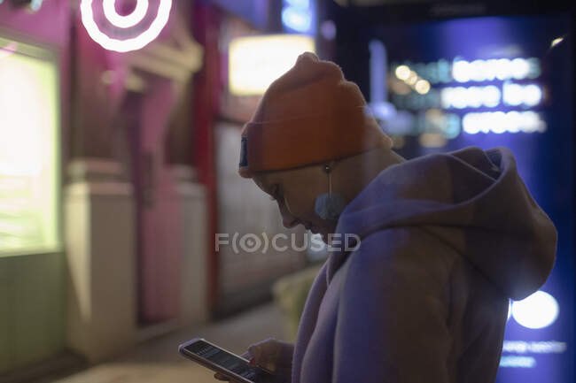 Mujer con estilo utilizando el teléfono inteligente en la acera de la ciudad por la noche - foto de stock