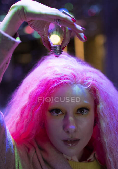 Portrait cool woman with pink hair holding light bulb over head — Stock Photo