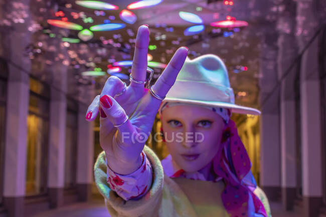 Portrait cool stylish woman in fedora gesturing peace sign under neon — Stock Photo