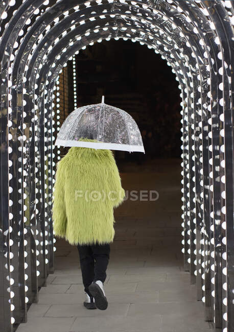 Eccentric young man in feather coat with umbrella under arch lights — Stock Photo