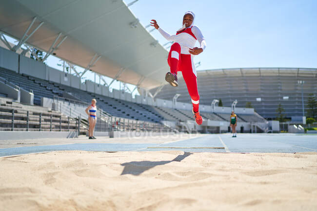 Atleta femminile di atletica leggera che salta lungo la sabbia nello stadio soleggiato — Foto stock