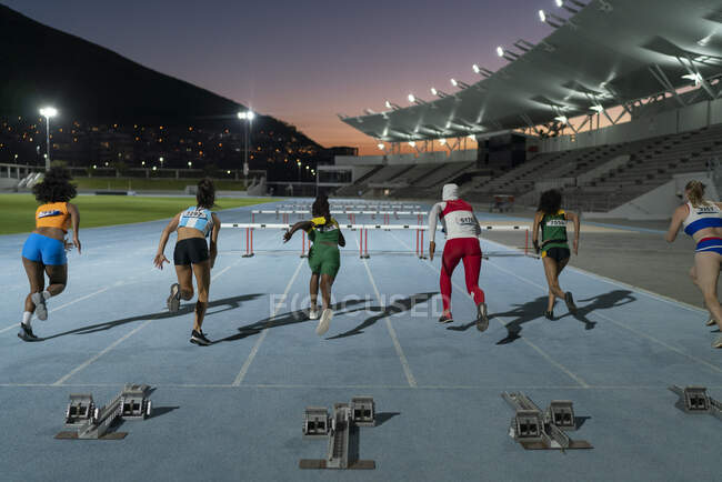 Female track and field athletes racing to hurdles on track at night — Stock Photo