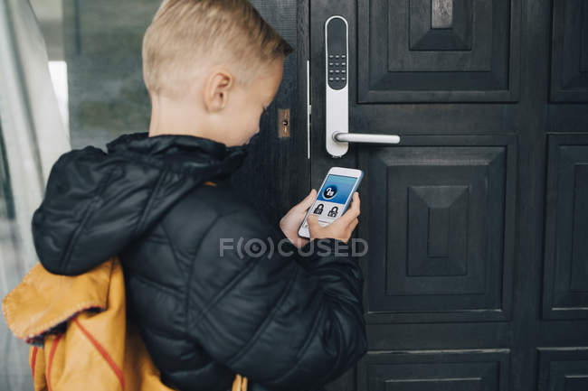 Rear View Of Boy Using App On Smart Phone To Unlock House Door