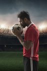 Jogador de futebol beijando futebol com estádio no fundo — Fotografia de Stock