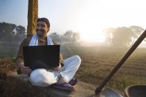 Agricultor sentado perto do campo agrícola e usando computador portátil — Fotografia de Stock