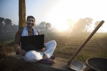 Фермер сидит рядом с сельскохозяйственным полем и использует ноутбук — стоковое фото