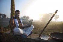 Agricultor sentado cerca del campo de la agricultura y el uso de ordenador portátil - foto de stock