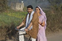 Happy rural couple in traditional dress riding on bicycle at country road — Stock Photo