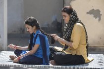 Женщина плетет волосы дочери сидя на раскладушке в доме — стоковое фото