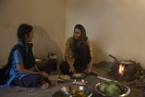 Mujer rural india sentada en la cocina y cocinando sobre leña con utensilios y verduras en el suelo y hablando con su hija - foto de stock