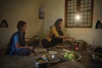 Indian rural woman sitting in kitchen and cooking on firewood with utensils and vegetables on floor and talking to daughter — Stock Photo