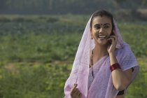 Retrato de mujer rural sonriente cubriendo la cabeza con sari hablando por teléfono contra el campo de la granja - foto de stock