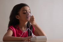 Young girl sitting and inhaling through a nebuliser at home. — Stock Photo
