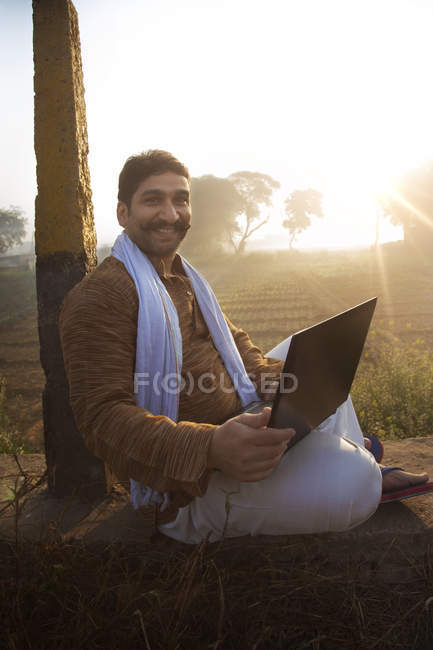 Фермер сидит рядом с сельскохозяйственным полем и использует ноутбук — стоковое фото