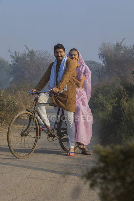 Happy rural couple in traditional dress riding on bicycle at country road — Stock Photo