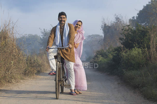 Feliz pareja rural en traje tradicional montando en bicicleta en el camino del campo - foto de stock