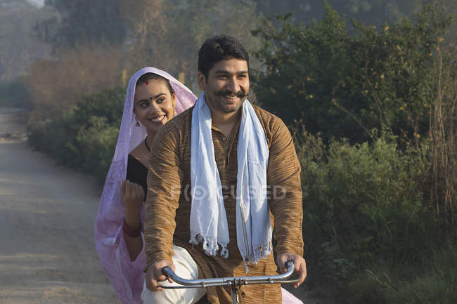 Feliz pareja rural en traje tradicional montando en bicicleta en el camino del campo - foto de stock