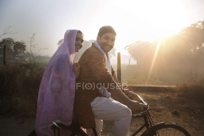 Vista laterale di felice coppia rurale in abito tradizionale in bicicletta su strada di campagna — Foto stock