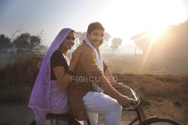 Vista lateral de la feliz pareja rural en traje tradicional a caballo en bicicleta en el camino del campo - foto de stock