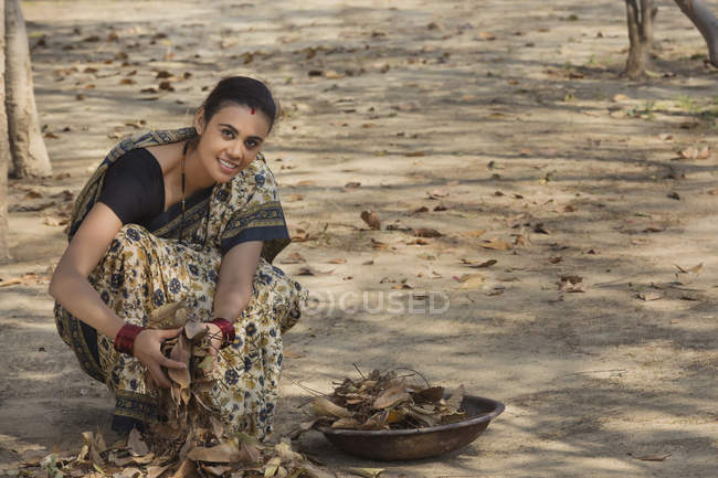 Indian woman dressed in sari collecting dried leaves from ground in iron gold pan — Stock Photo