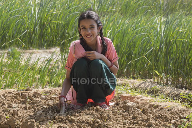 Маленькая индийская девочка копает землю с помощью лопатки сидя в сельском хозяйстве — стоковое фото