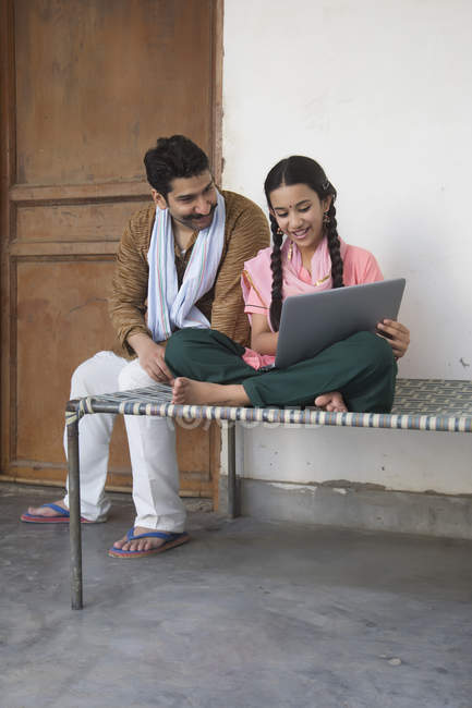 Girl sitting with father on cot and using laptop computer — Stock Photo