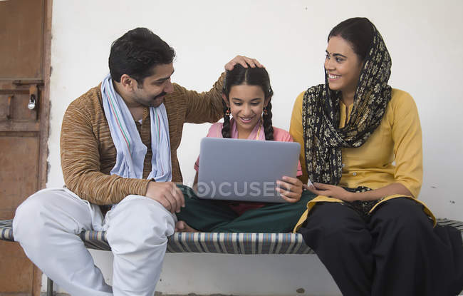 Индийская девушка сидит с родителями на детской кроватке и с помощью ноутбука — стоковое фото
