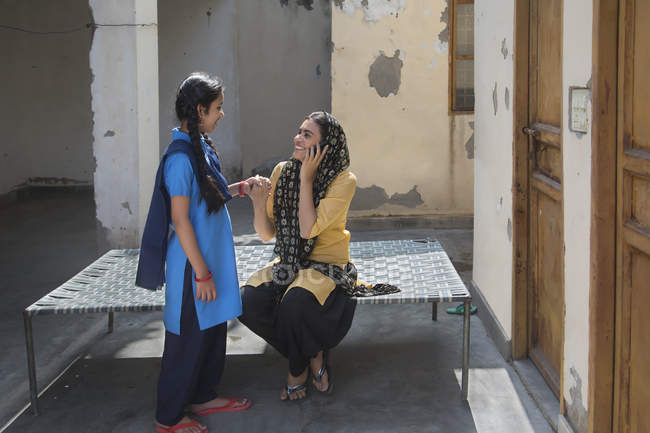 Индийская девушка рядом с молодой взрослой женщиной разговаривает по мобильному телефону, сидя на койке — стоковое фото