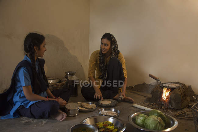 Indian rural woman sitting in kitchen and cooking on firewood with utensils and vegetables on floor and talking to daughter — Stock Photo
