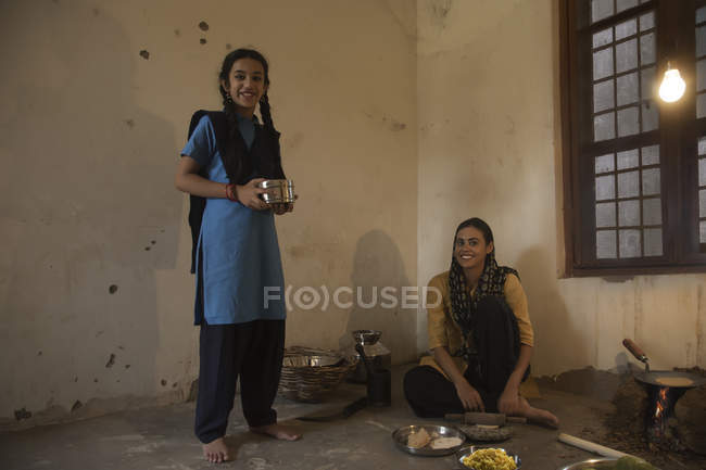 Indische Landfrau sitzt in Küche auf dem Boden, während Tochter mit Tiffinbox steht — Stockfoto