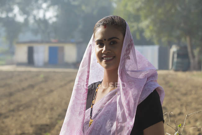 Retrato de mujer rural sonriente cubriendo la cabeza con sari sentado en el campo - foto de stock