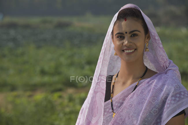 Retrato de mujer rural sonriente cubriendo la cabeza con sari contra el campo agrícola - foto de stock