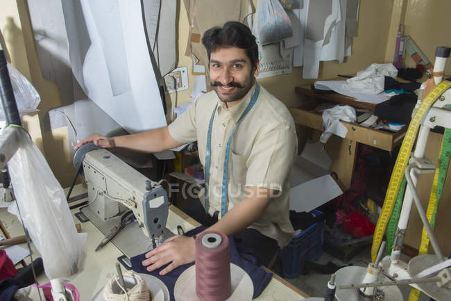 Alfaiate masculino trabalhando na máquina de costura na oficina com acessórios de alfaiataria pendurados ao redor — Fotografia de Stock